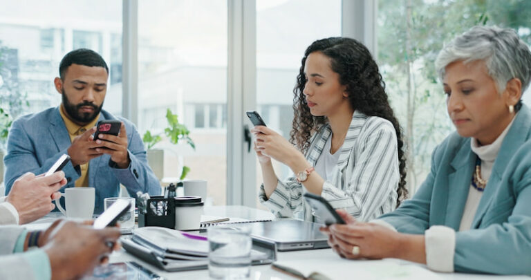 Vier Beschäftigte sitzen im Meeting am Tisch und schauen auf ihr Smartphone, dem Produktivitätskiller, anstatt sich zu unterhalten und zu arbeiten.