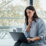 Eine Arbeitnehmerin sitzt auf dem Sofa und schaut nachdenklich auf ihren Laptop. Sie überlegt, wie gut sie ihren Arbeitgeber bewerten möchte. Dafür nutzt sie eine Bewertungsplattform.