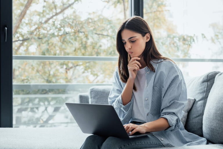 Eine Arbeitnehmerin sitzt auf dem Sofa und schaut nachdenklich auf ihren Laptop. Sie überlegt, wie gut sie ihren Arbeitgeber bewerten möchte. Dafür nutzt sie eine Bewertungsplattform.