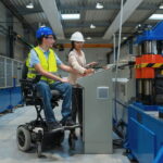 Eine Vorgesetzte erklärt einem Arbeitnehmer im Rollstuhl die Bedienung einer Maschine. Er durchläuft eine berufliche Reha in Form einer Umschulung.
