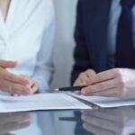 Ein Chef im Anzug geht einen Änderungsvertrag zum Arbeitsvertrag mit einer Mitarbeiterin durch. Er zeigt mit einem Kugelschreiber auf den Vertrag, während die Angestellte mit ihrer Hand gestikuliert.