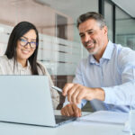 Ein Vorgesetzter sitzt mit einer neuen Mitarbeiterin vor einem Laptop und zeigt ihr Aufgaben, die in ihrer Arbeitsplatzbeschreibung genannt werden.