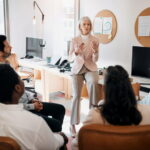 Eine Businessfrau steht vor einer Gruppe Arbeitnehmer und hält einen Vortrag über Compliance im Unternehmen.