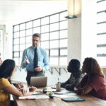 Eine männliche Führungskraft im Anzug steht im Meeting vor ihrem Team und gibt Anweisungen. Das Team besteht aus vier Personen, die an einem Tisch sitzen. Die Leitungsspanne ist klein.