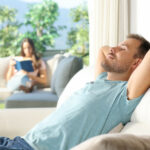 Ein Mann entspannt sich draußen auf einem Gartensofa und entspannt sich. Im Hintergrund ist eine Frau sichtbar, die ein Buch liest. Sie wollen abschalten nach der Arbeit.
